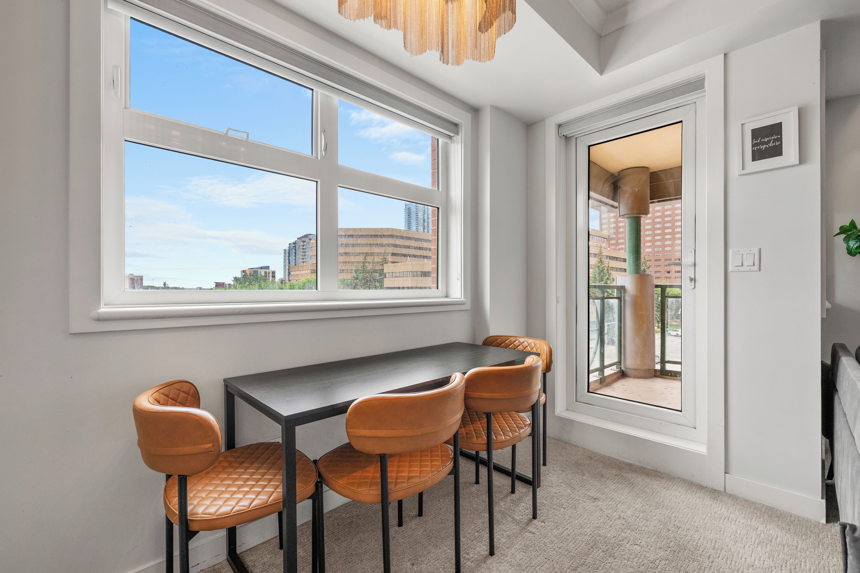 Una habitación con una mesa de comedor y cuatro sillas, ventanas amplias con vistas a edificios y una puerta que da a un área exterior.