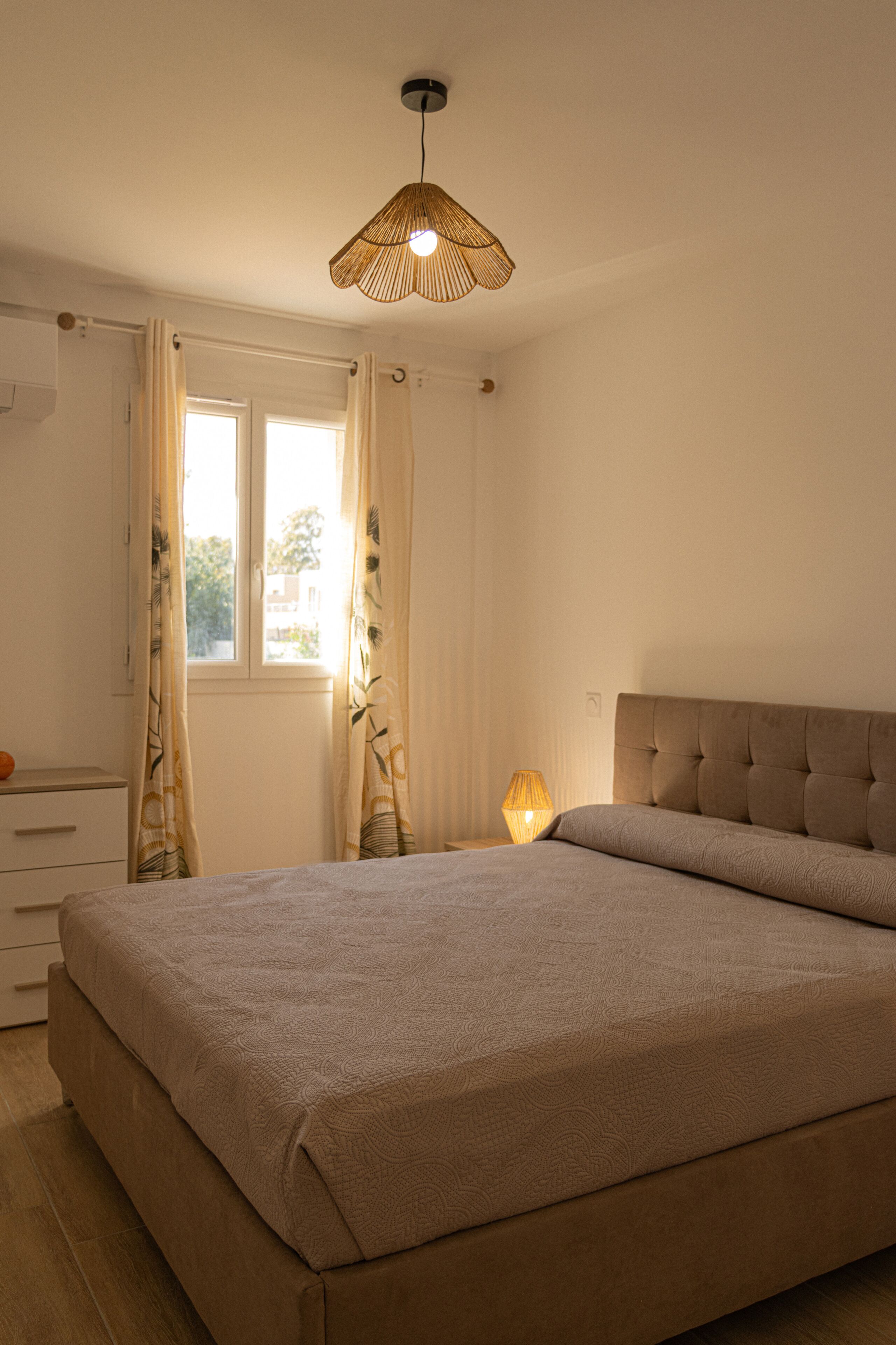 Un dormitorio con cama, lámpara de noche, ventana con cortinas y una lámpara colgante.
