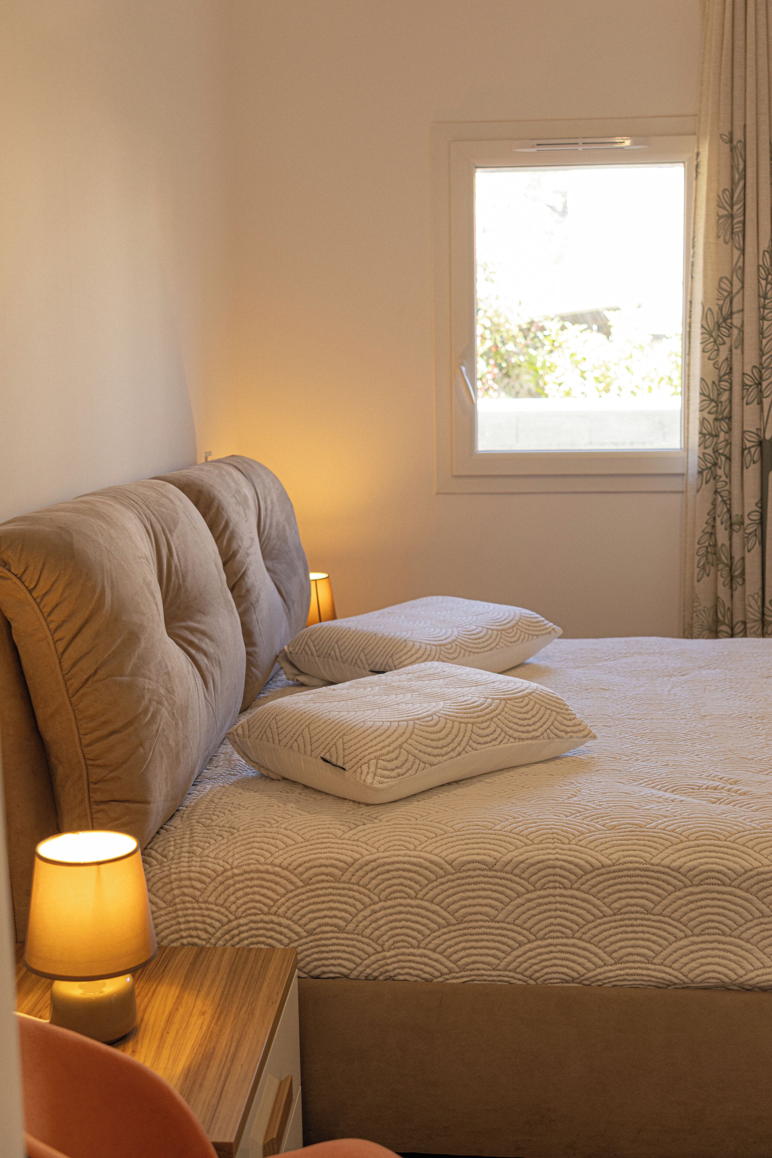 Un dormitorio con una cama, almohadas, una mesita de noche con lámpara, una ventana y cortinas.