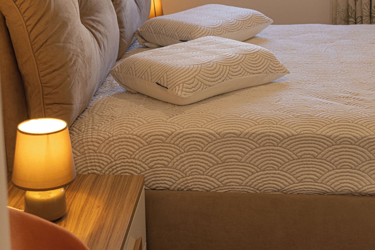 A bedroom with a bed, pillows, a nightstand with a lamp, a window, and curtains.