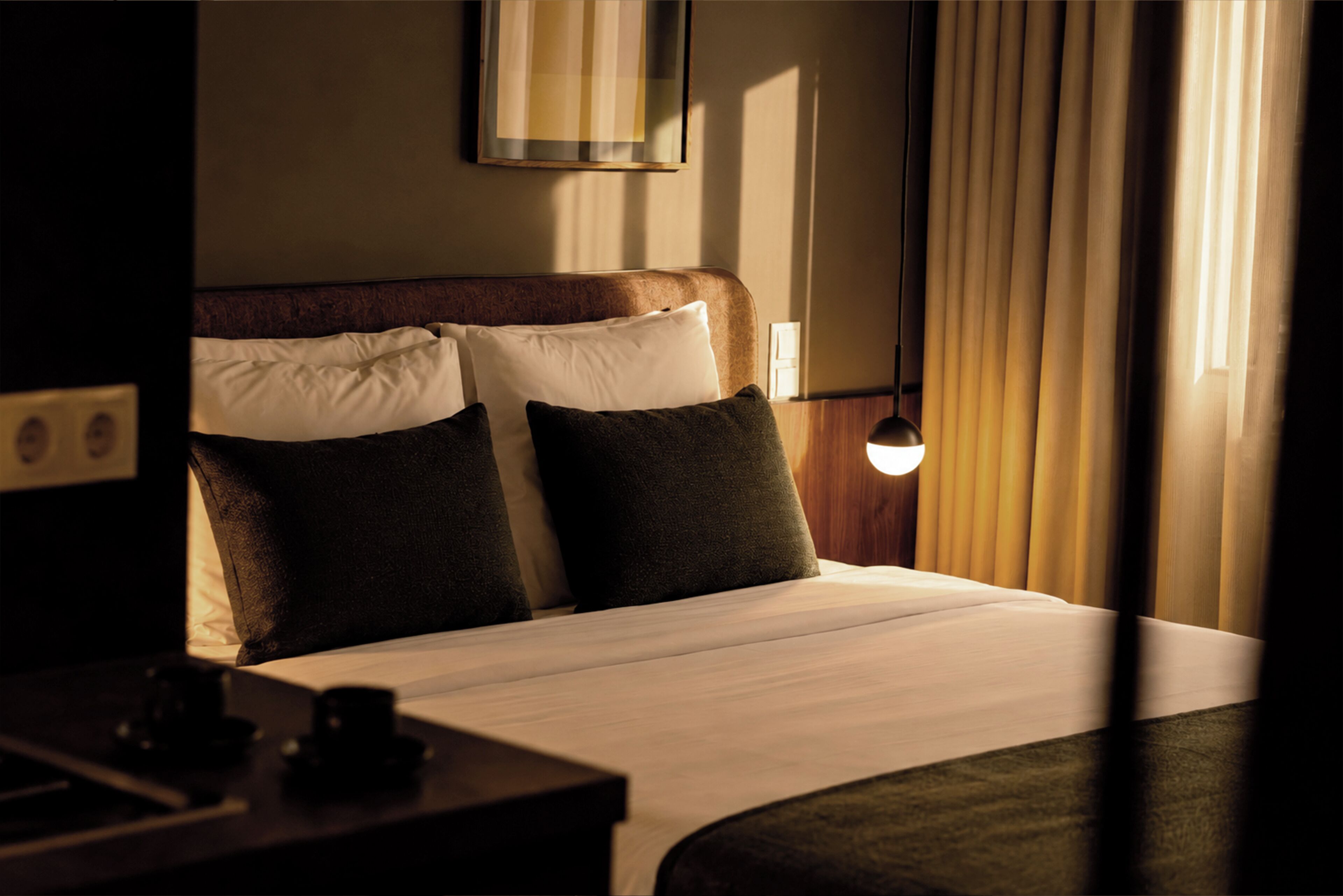 Hotelkamer met een bed, kussens, een nachtkastje, een lamp en een raam met gordijnen.