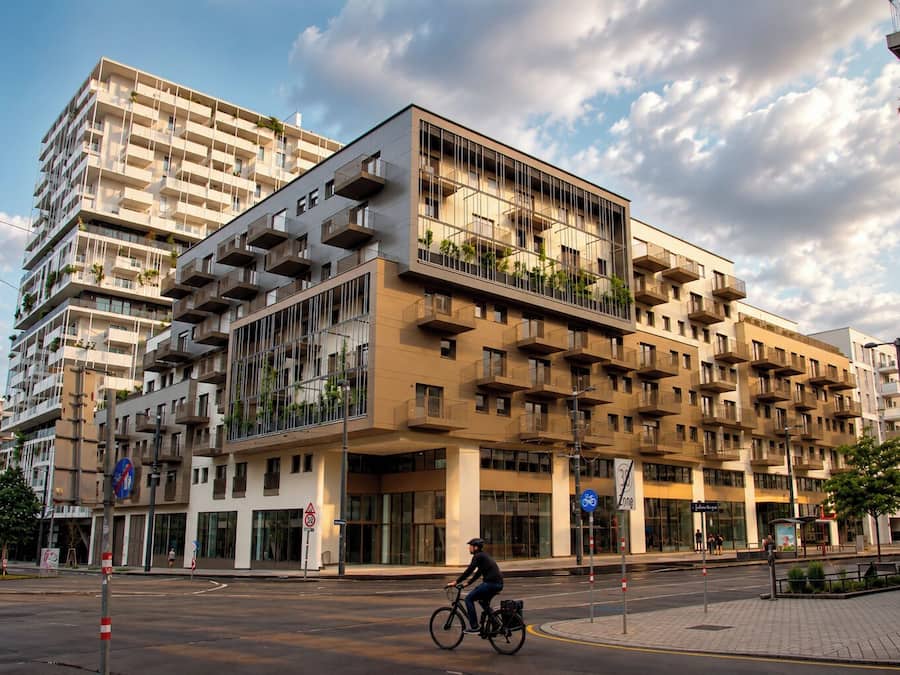 A multi-story residential building with balconies, a cyclist on the street, and a clear sky.