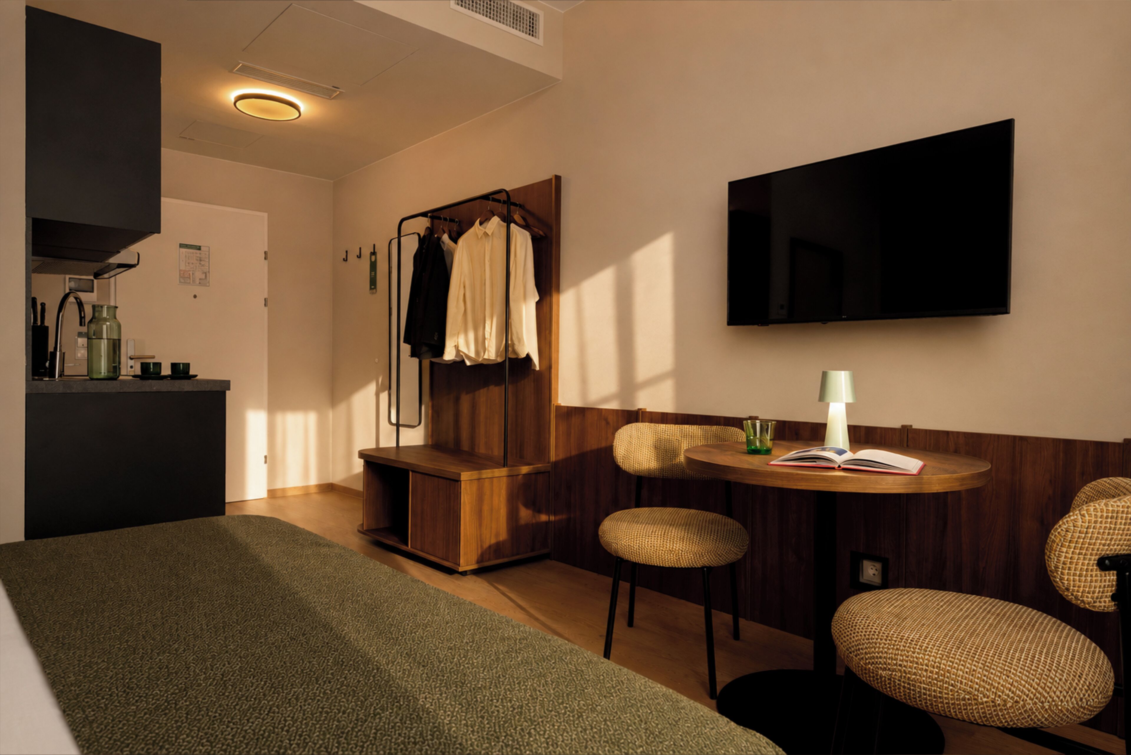 Una habitación de hotel moderna con una cama, una zona de estar con una mesa y sillas, un televisor montado en la pared y un armario con ropa colgada.