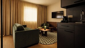 A modern hotel room with a sofa, a small table with a lamp, a TV mounted on the wall, and a kitchenette area. - Habyt Vienna (Vienna)