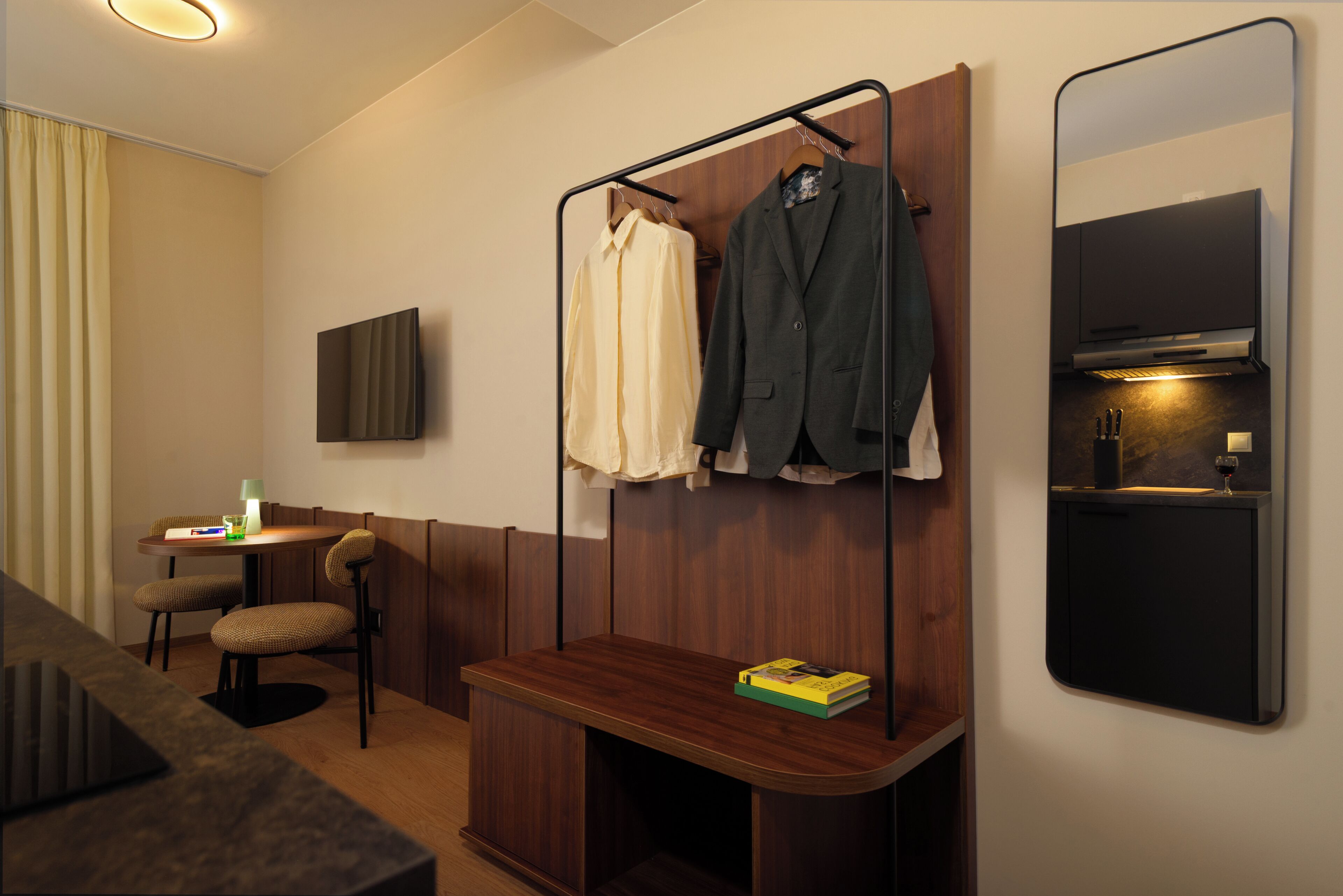 Habitación de hotel con un banco de madera, una mesita con lámpara, un televisor montado en la pared y un perchero con dos chaquetas.