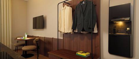 Habitación de hotel con un banco de madera, una mesita con lámpara, un televisor montado en la pared y un perchero con dos chaquetas.