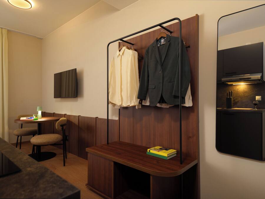 A hotel room with a wooden bench, a small table with a lamp, a TV mounted on the wall, and a coat rack with two jackets.
