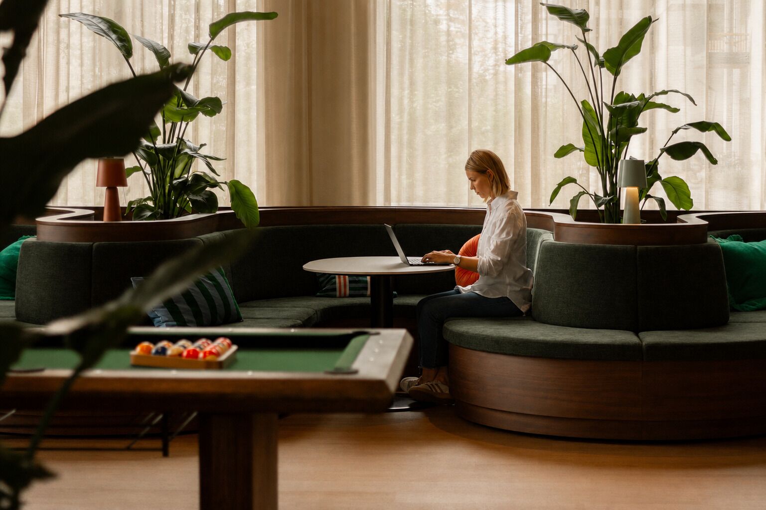 A person seated on a curved sofa with a laptop, surrounded by potted plants and a pool table in a room with large windows.