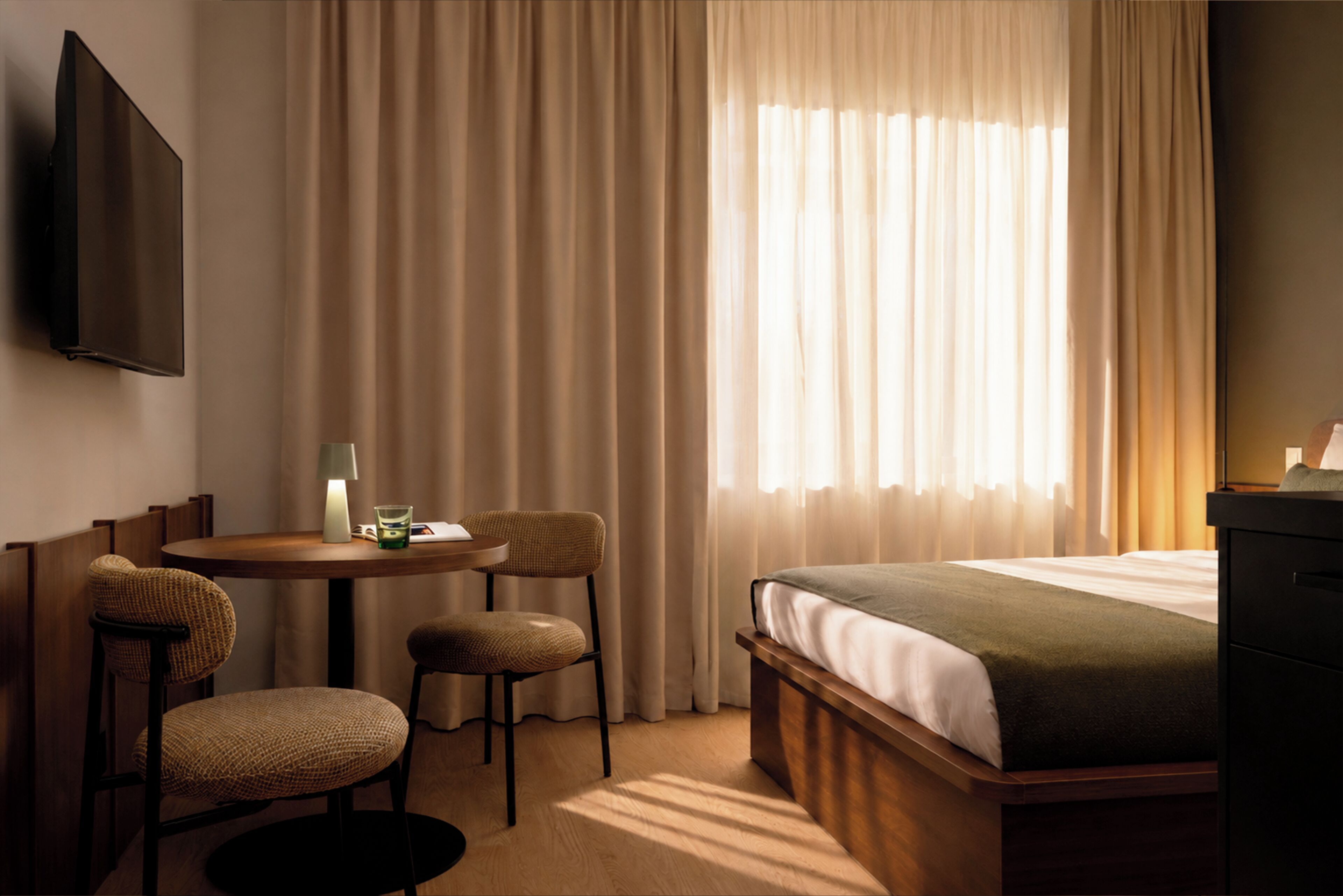 Quarto de hotel com cama, escrivaninha com duas cadeiras, televisão na parede e janela com cortinas.