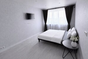 Moderni hotellihuone, jossa on sänky, taulutelevisio, ikkuna verhoilla ja pieni pöytä, jonka päällä on maljakko täynnä kukkia.