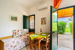 Ein Zimmer mit einem Bett, einem Esstisch mit Stühlen, einer offenen Tür zu einem Garten und einer Klimaanlage an der Wand.