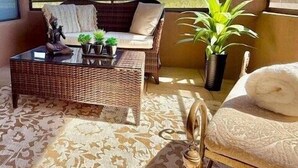 Un balcón con un sofá de mimbre, una mesita con plantas, una planta en maceta y una silla con una toalla.