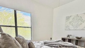 Un dormitorio con un ventanal grande, una cama con ropa de cama estampada, una mesita de noche con un objeto decorativo y una obra de arte colgada en la pared.