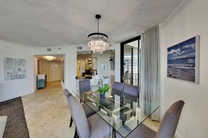 Ein moderner Essbereich mit einem Glastisch, grauen Stühlen, einer Kronleuchter und Blick auf das Meer durch ein großes Fenster.