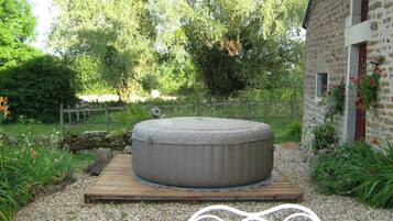 A hot tub on a wooden platform in a garden with a stone building and a wooden fence in the background.