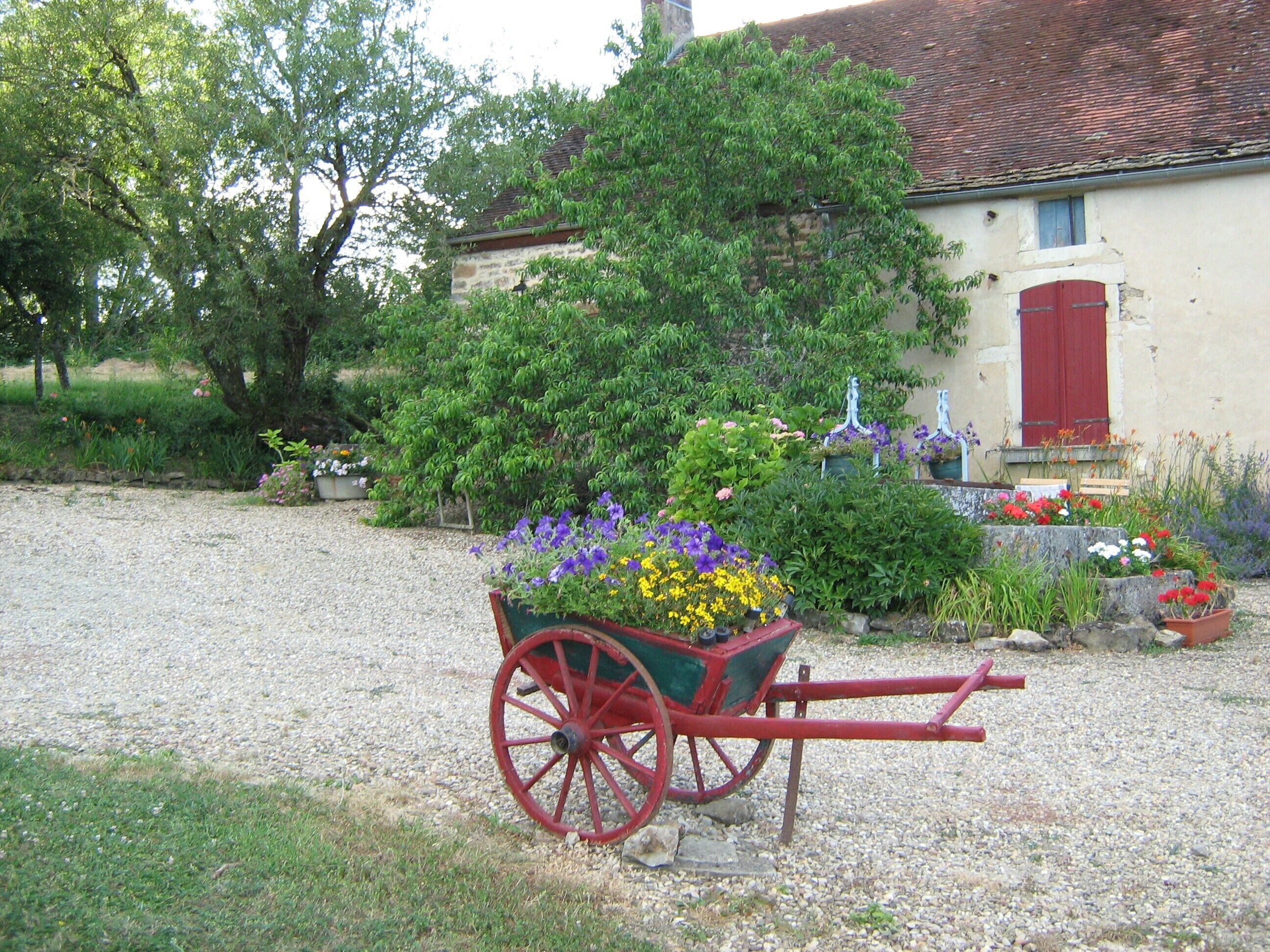 Ein Kiesweg führt zu einem Haus mit roter Tür und einer Schubkarre voller Blumen.