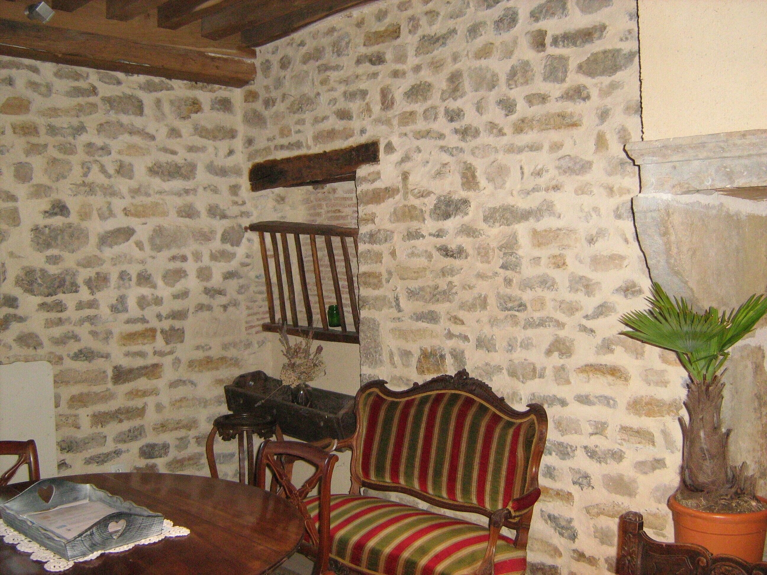 Une pièce aux murs de pierre, avec une table à manger en bois, une chaise à rayures, une plante en pot et une étagère sur laquelle se trouve une bouteille.