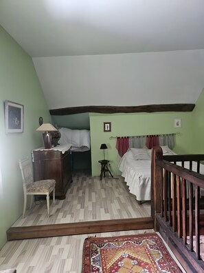 A room with a wooden floor, a bed with white bedding, a chair, a small table with a lamp, and a picture frame on the wall.
