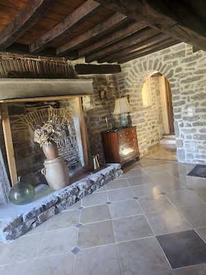 Une cheminée en pierre ornée d’un vase de fleurs séchées, un meuble en bois et un sol carrelé.