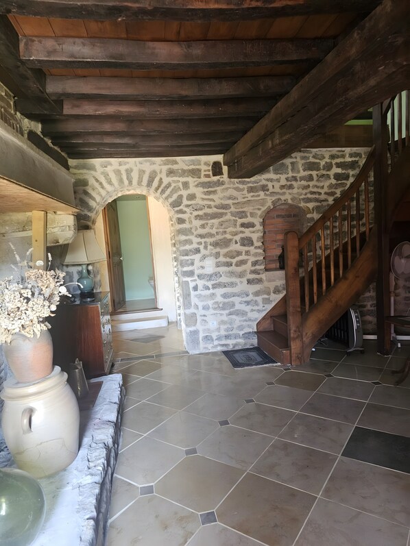 A stone-walled interior with a wooden staircase, a tiled floor, and a vase with dried flowers on a table.