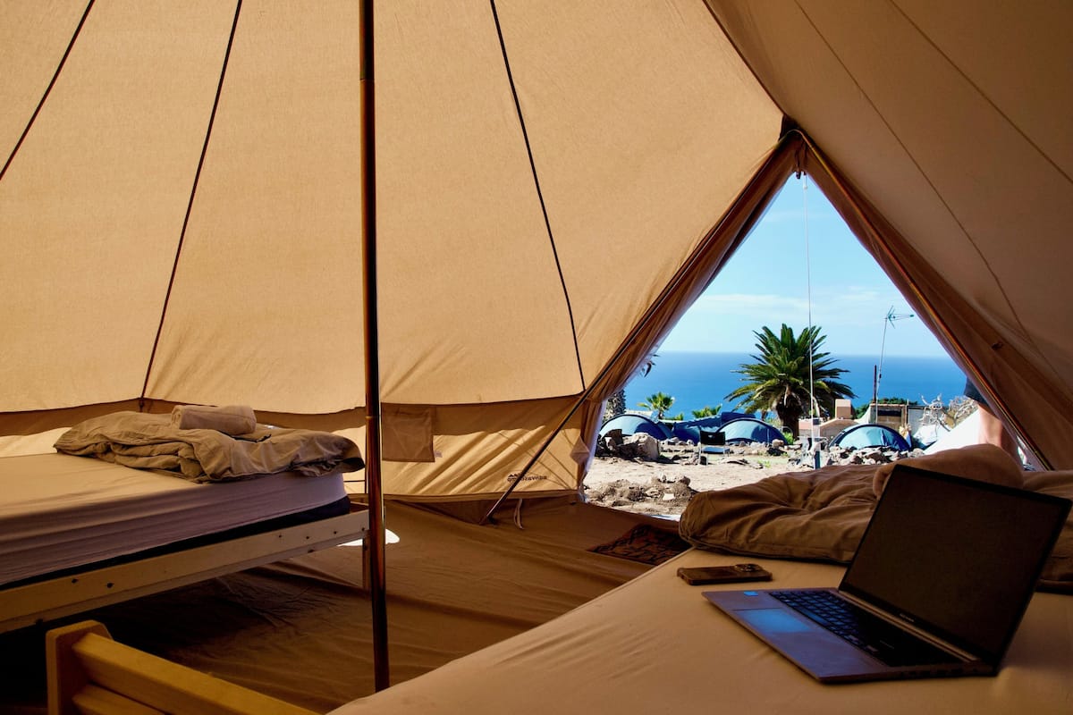 A tent with a bed and a laptop, a window with a view of the beach and tents.