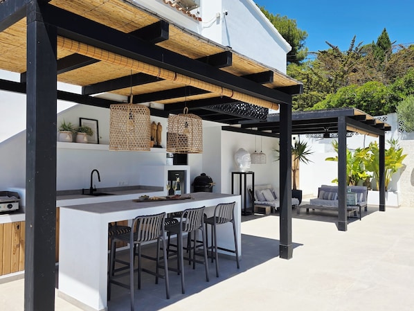 An outdoor kitchen with a white countertop, black bar stools, and hanging wicker light fixtures.