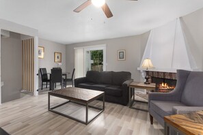 Un salon avec une cheminée, un ventilateur de plafond, un coin repas avec des chaises et un canapé.