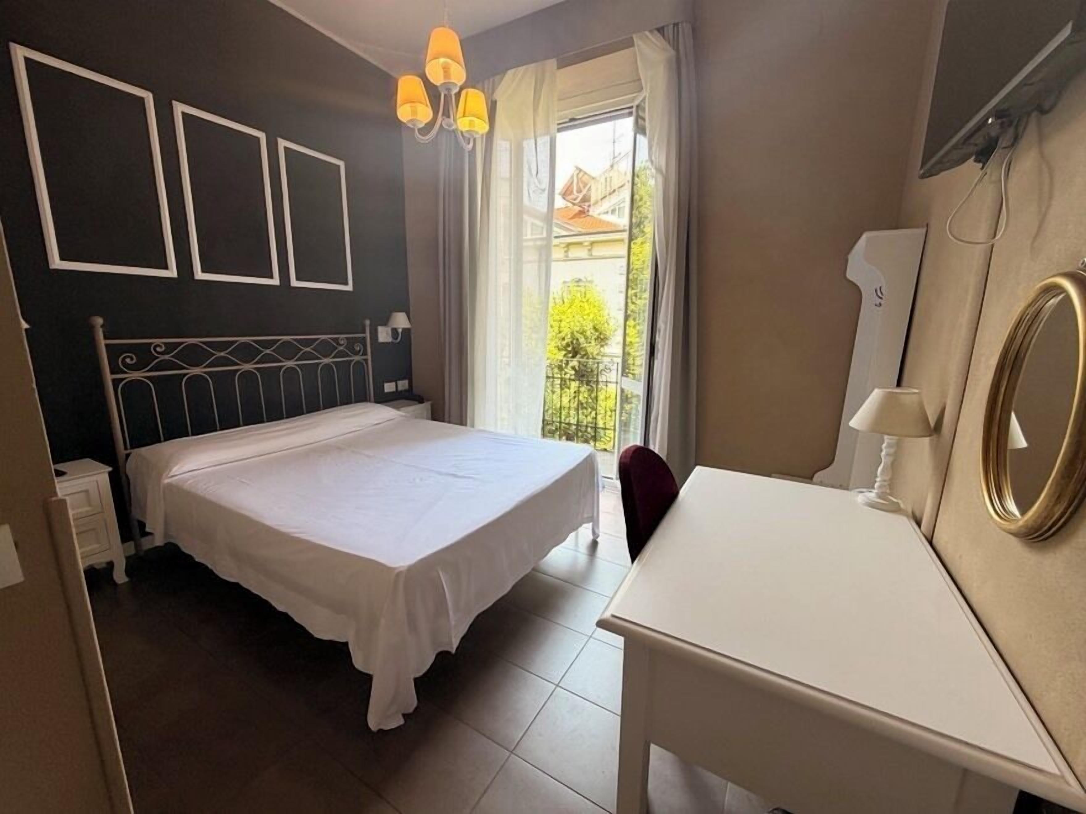 A bedroom with a bed, a desk, and a chair. There is a window with curtains, a chandelier, and a mirror on the wall.