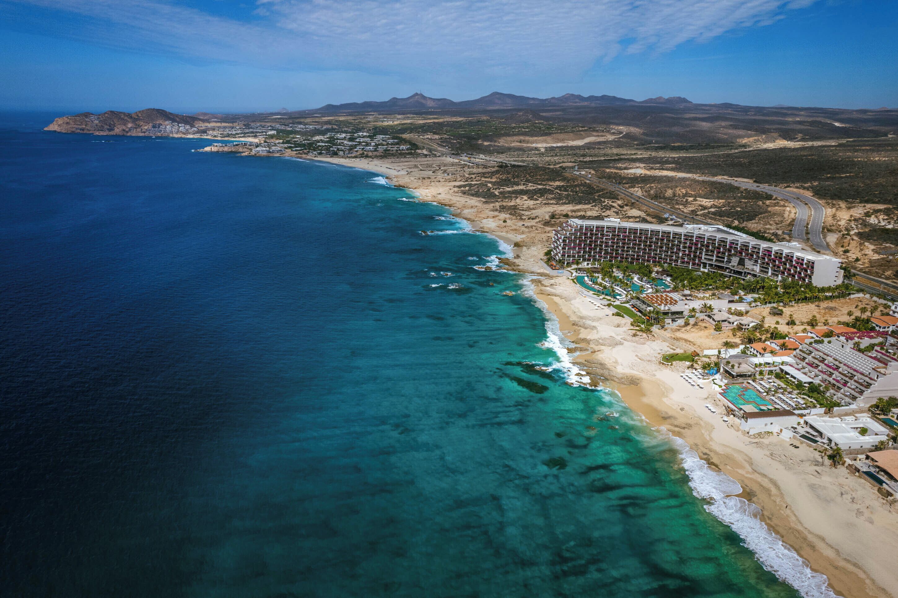An aerial view of a coastal hotel with a beach, clear blue waters, and mountainous background.