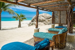 Eine Strandlounge mit Korbmöbeln und Kissen mit Blick auf einen Sandstrand und den Ozean.