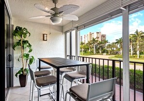 Ein Balkon mit Tisch und Stühlen, einem Deckenventilator, einem Blumentopf und Blick auf Palmen und Gebäude.