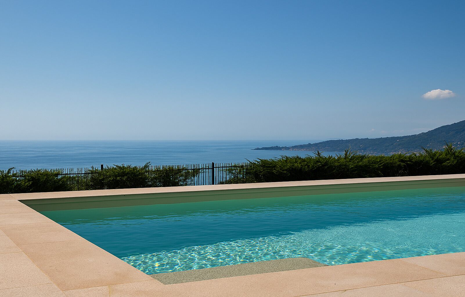A rectangular swimming pool with clear blue water, surrounded by a beige deck, and a view of the sea and distant hills.