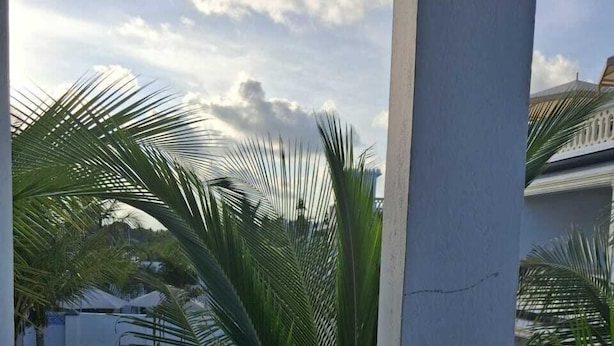A balcony with a white railing, palm trees, and a building with blue roofing in the background. - Makey Holiday Homes Diani (Diani Beach)
