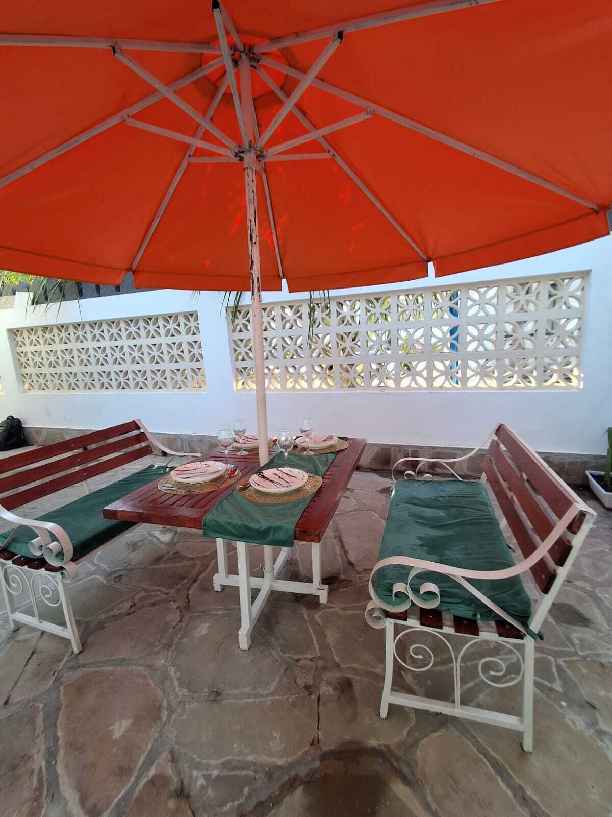 An outdoor patio with stone flooring, wooden benches, a table set for dining, and an orange umbrella.