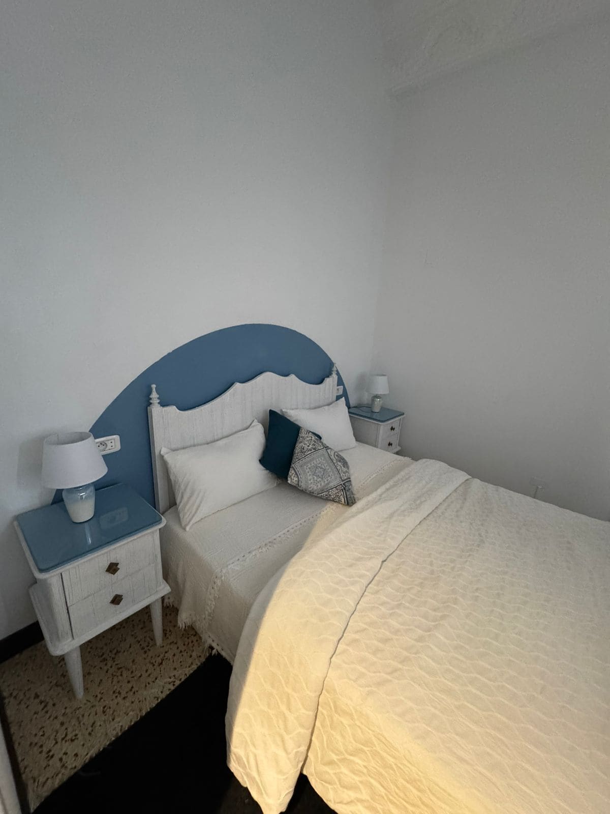 Un dormitorio con cama, mesita de noche, lámpara y una pequeña mesa junto a la cama.