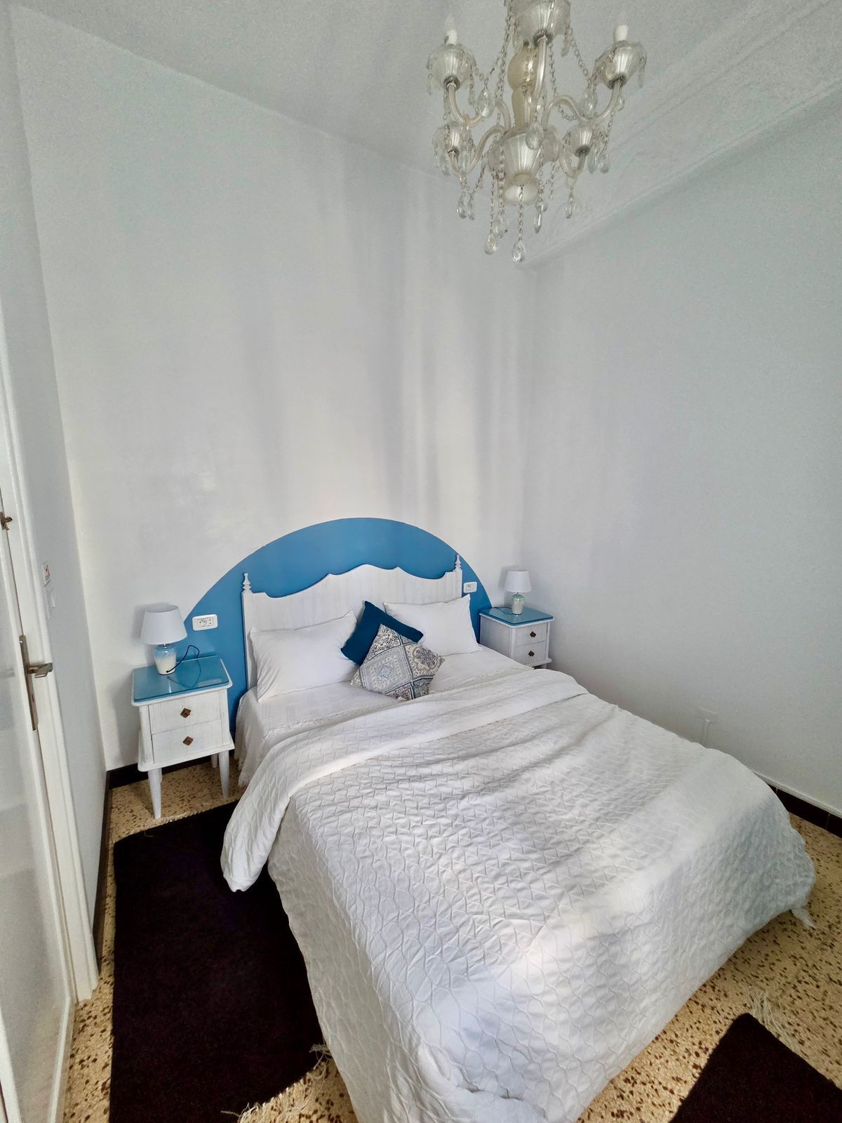 A bedroom with a bed, a chandelier, two bedside tables with lamps, and a door.