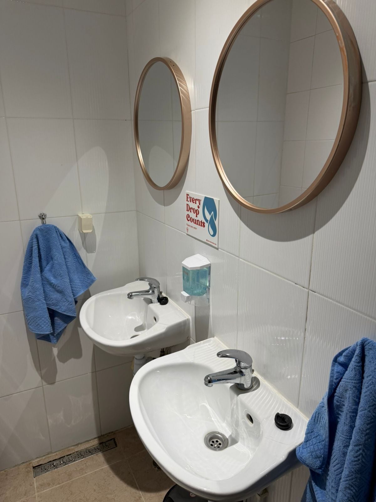 Baño con dos lavamanos, una toalla azul, dispensador de jabón y un cartel que dice 'Cada gota cuenta'.