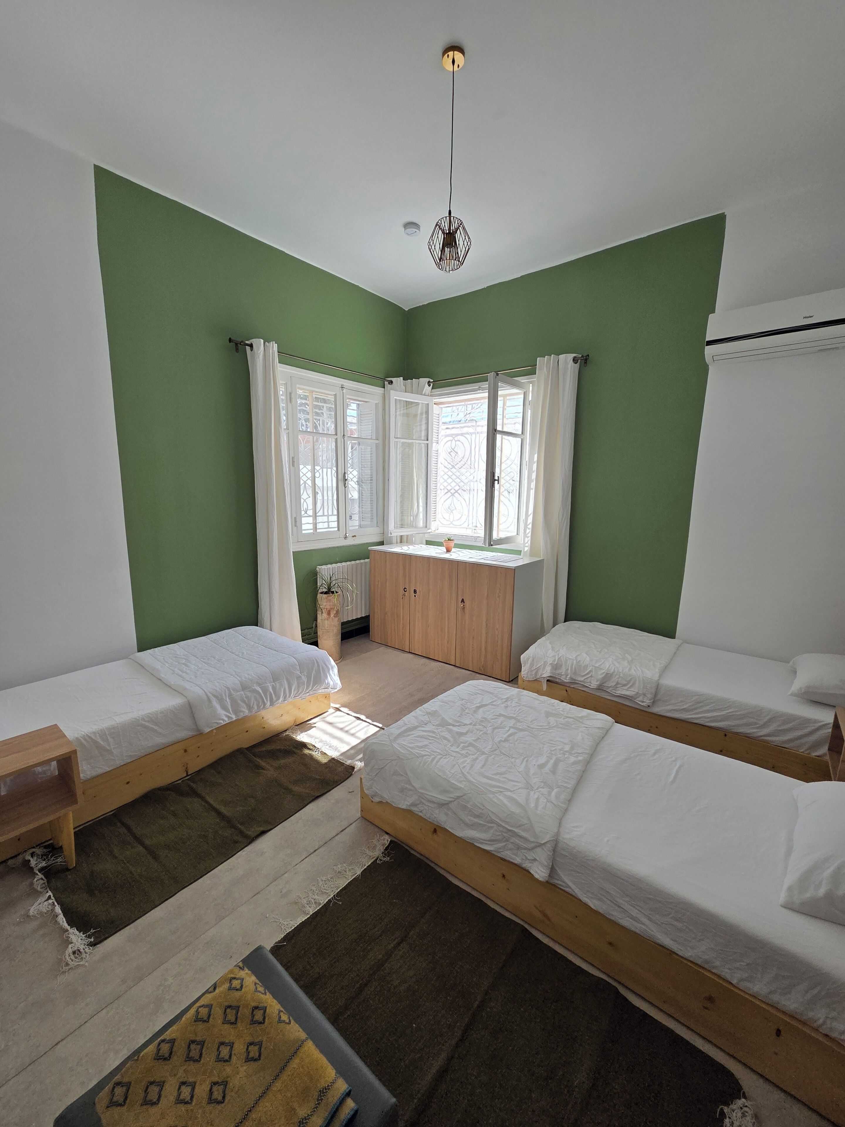 Una habitación con tres camas de madera, una ventana con cortinas blancas, un radiador y una lámpara colgante.