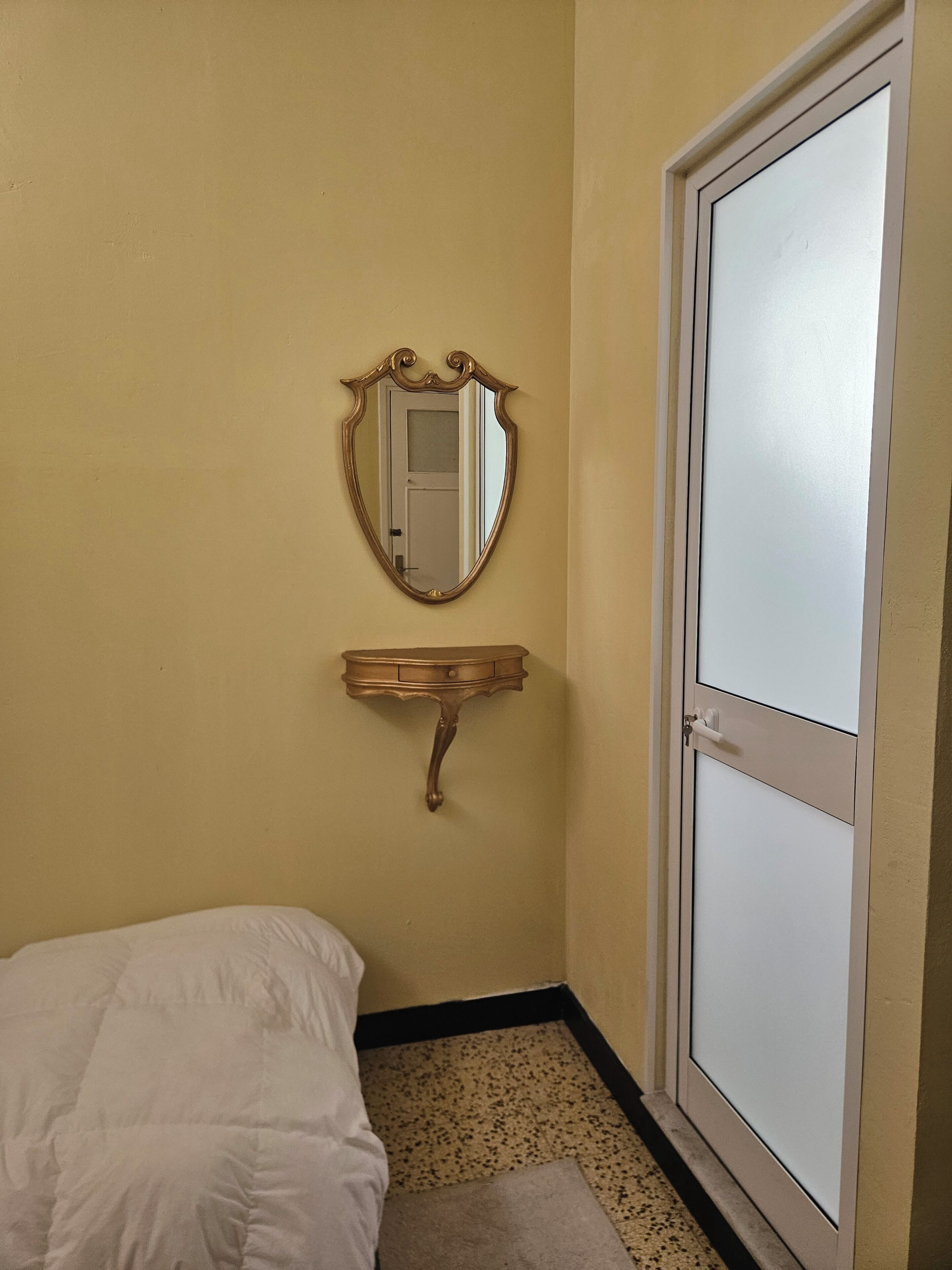 Quarto de hotel pequeno e individual, com uma cama, um espelho fixado na parede, uma cômoda de madeira e uma porta.