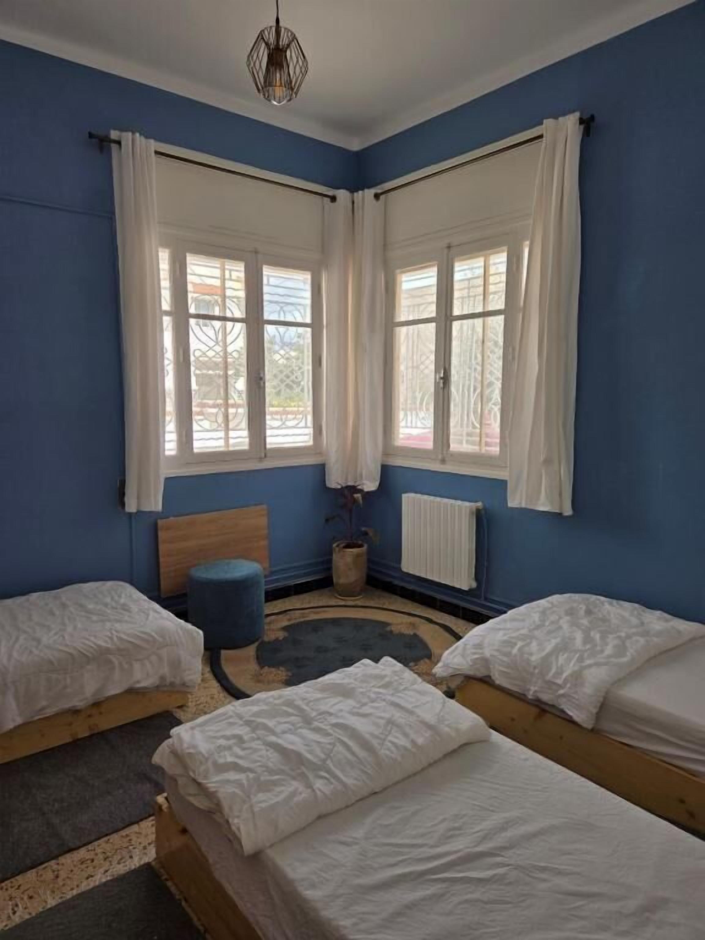 Een kamer met twee bedden, een blauwe wand, een houten hoofdbord, een blauwe poef, een radiator en een hanglamp.