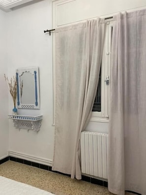 Un baño con un radiador blanco, una ventana con cortina y un estante pequeño con un florero.