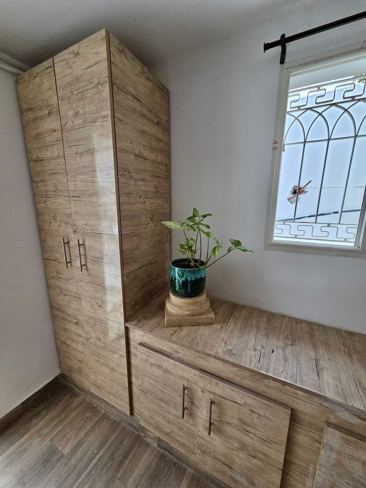 A room with a wooden wardrobe, a small bench with a potted plant, and a window with a metal grille.