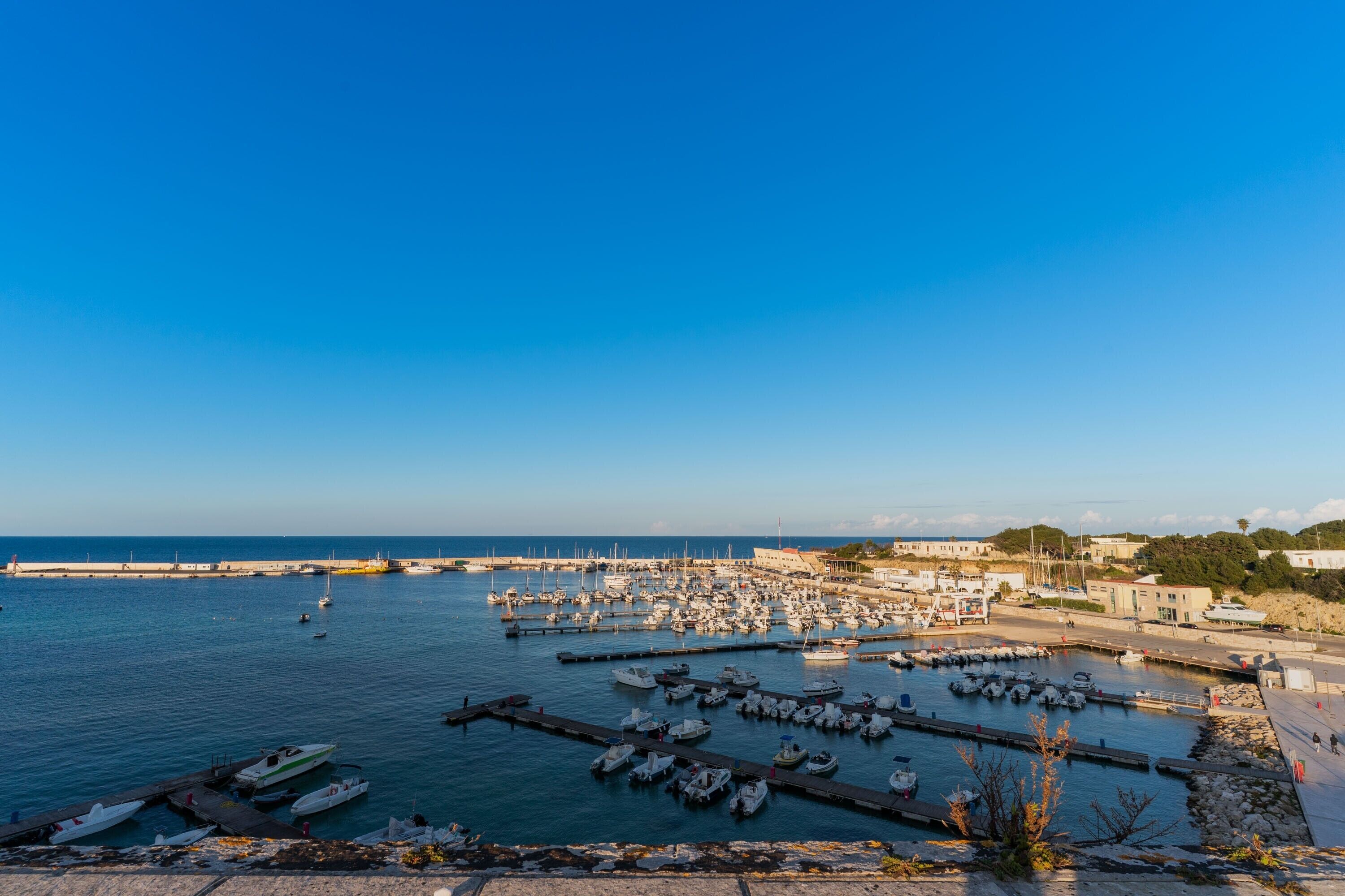 Uma marina com barcos atracados, um cais que se estende sobre a água e edifícios ao fundo.