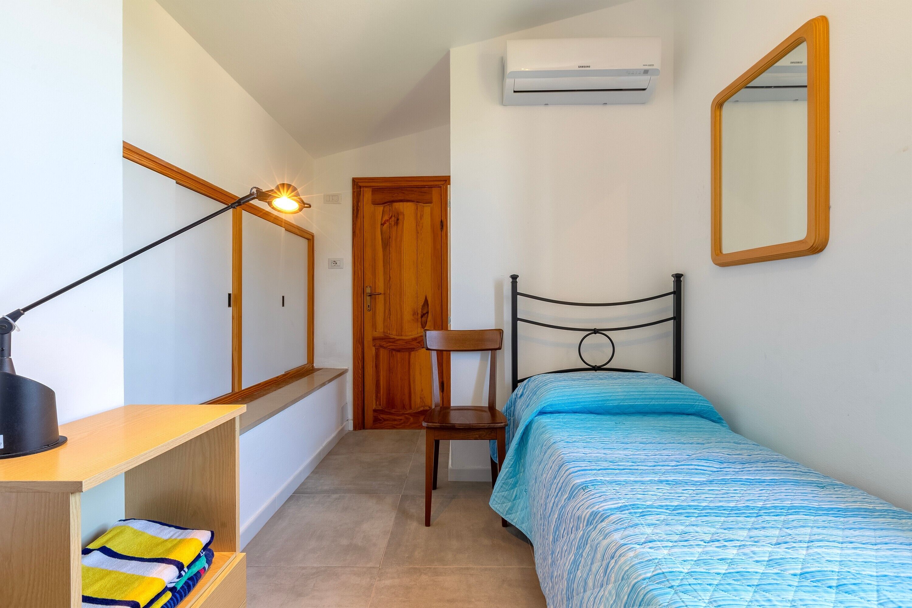 Ein Schlafzimmer mit einem Bett, einem Sessel, einem Schreibtisch mit Lampe, einem Spiegel und einer Klimaanlage.