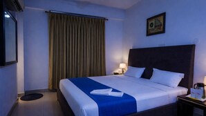 Habitación de hotel con una cama grande, una mesita de noche con lámpara, un cuadro enmarcado en la pared y una ventana con cortinas.