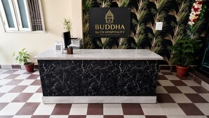 Un mostrador de recepción con encimera de mármol y base negra, flanqueado por plantas en macetas y un letrero que dice 'BUDDHA by CN HOSPITALITY'.