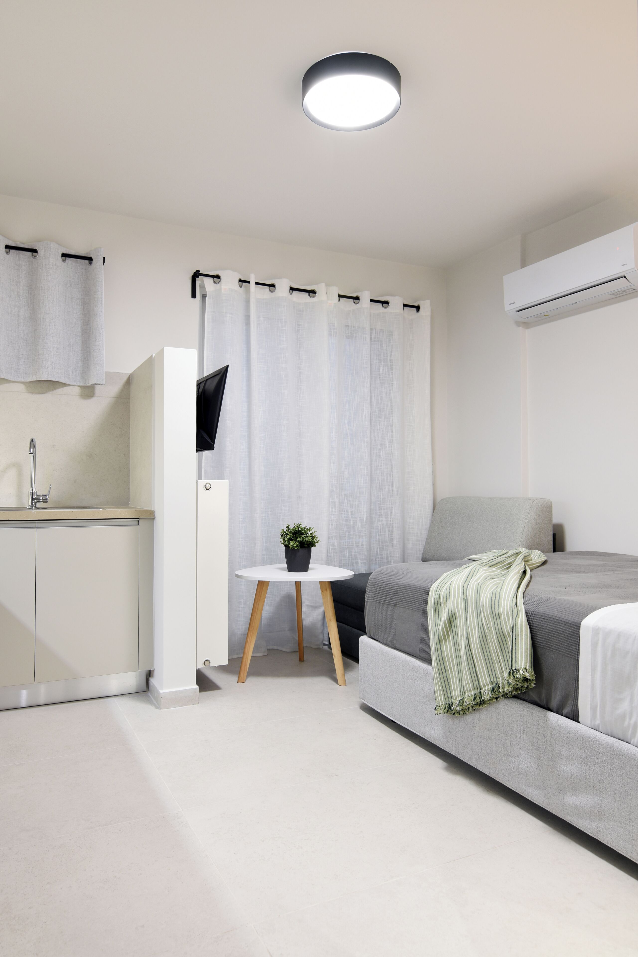 Ein modernes, minimalistisches Zimmer mit einem Bett, einem kleinen Tisch mit Pflanze, einem an der Wand montierten Fernseher und einer Küchenzeile.