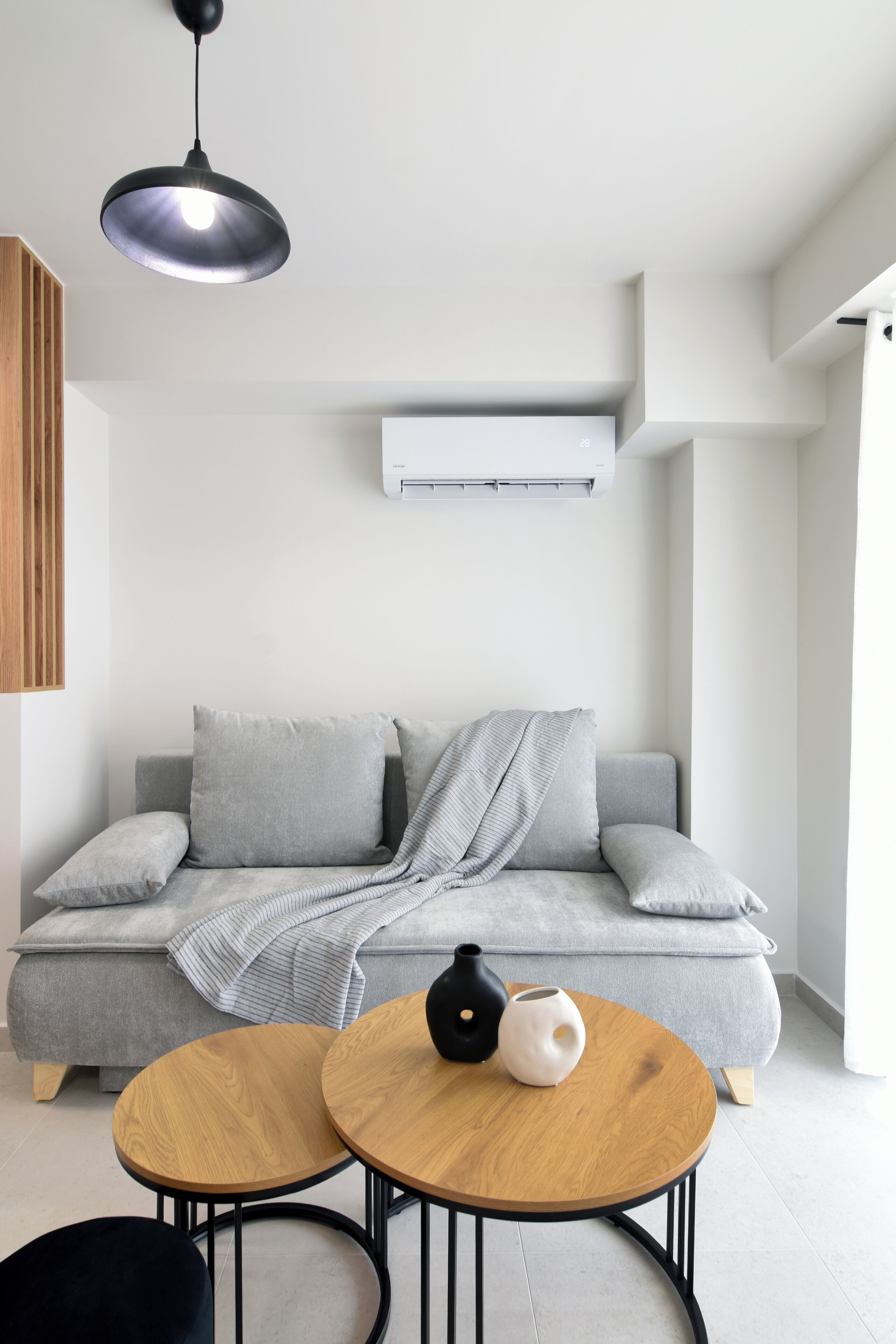 Un salon moderne avec un canapé gris, deux petites tables rondes en bois, un vase noir et blanc et une suspension.
