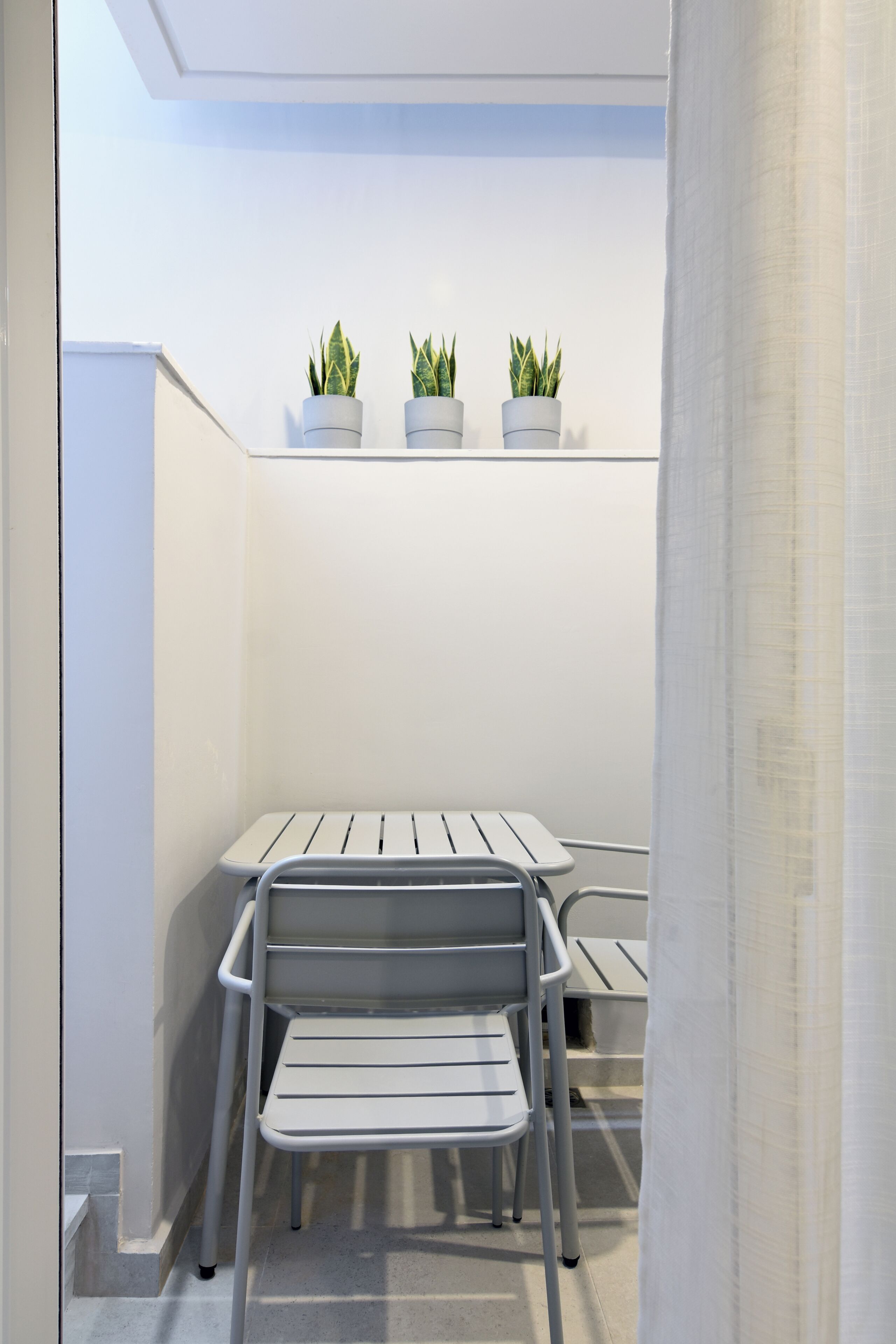 A small, enclosed balcony with a white table and two chairs, three potted plants on a shelf, and a white curtain.