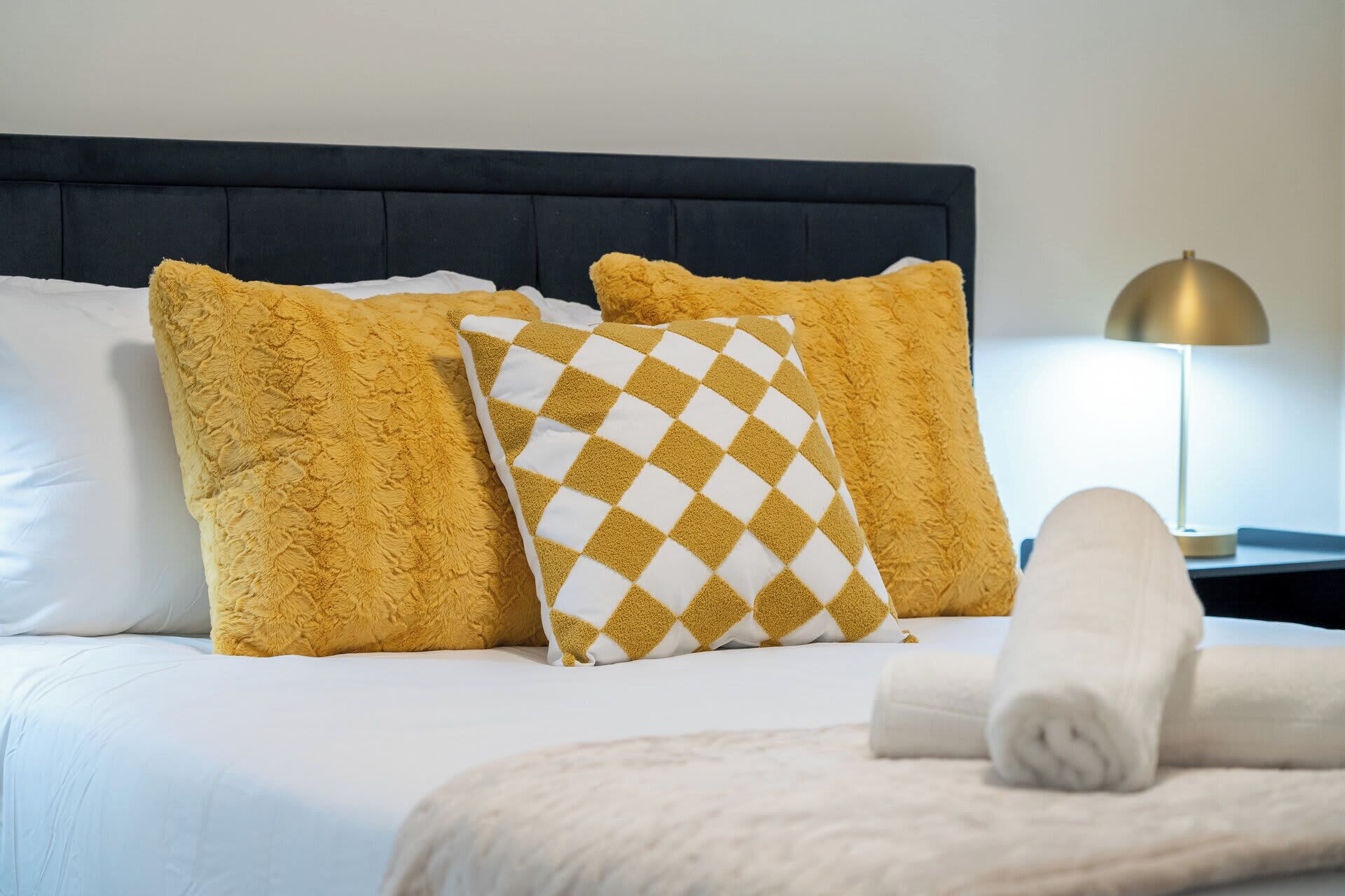 A neatly made bed with a dark headboard, two fluffy pillows, a checkered pillow, and a rolled towel.
